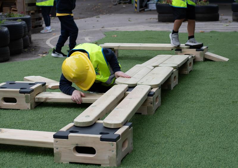 A child dressed in a hi-vis Pentagon Play jacket with a yellow hard hat is connecting a wooden plank to a wooden block from the Play Builder Architect set.